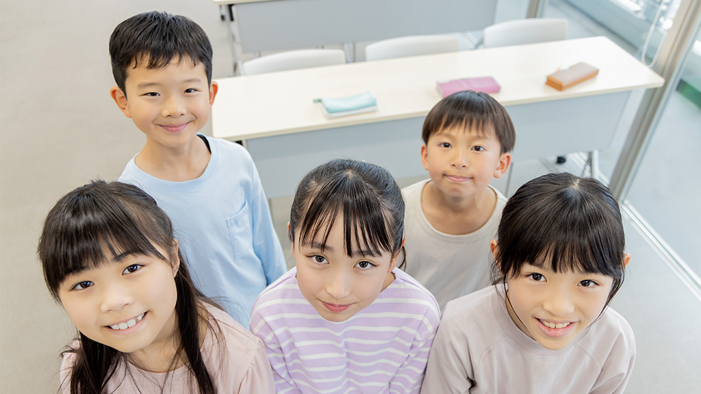 地域史を学ぶ市民参加型ワークショップの教育プログラムに参加する小学生たちが、教室内で楽しそうにカメラを見つめている場面のイメージ画像